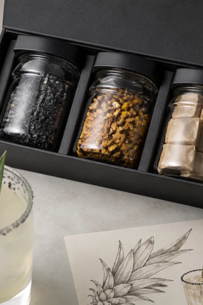 A set of three small jars in a matte black box. Jar 1 has gray-black smoked salt. Jar 2 has amber tonic mix with tiny dried pineapple bits. Jar 3 contains brown sugar cubes wrapped in parchment. A recipe card shows a smoky garnish with a pineapple leaf sketch.