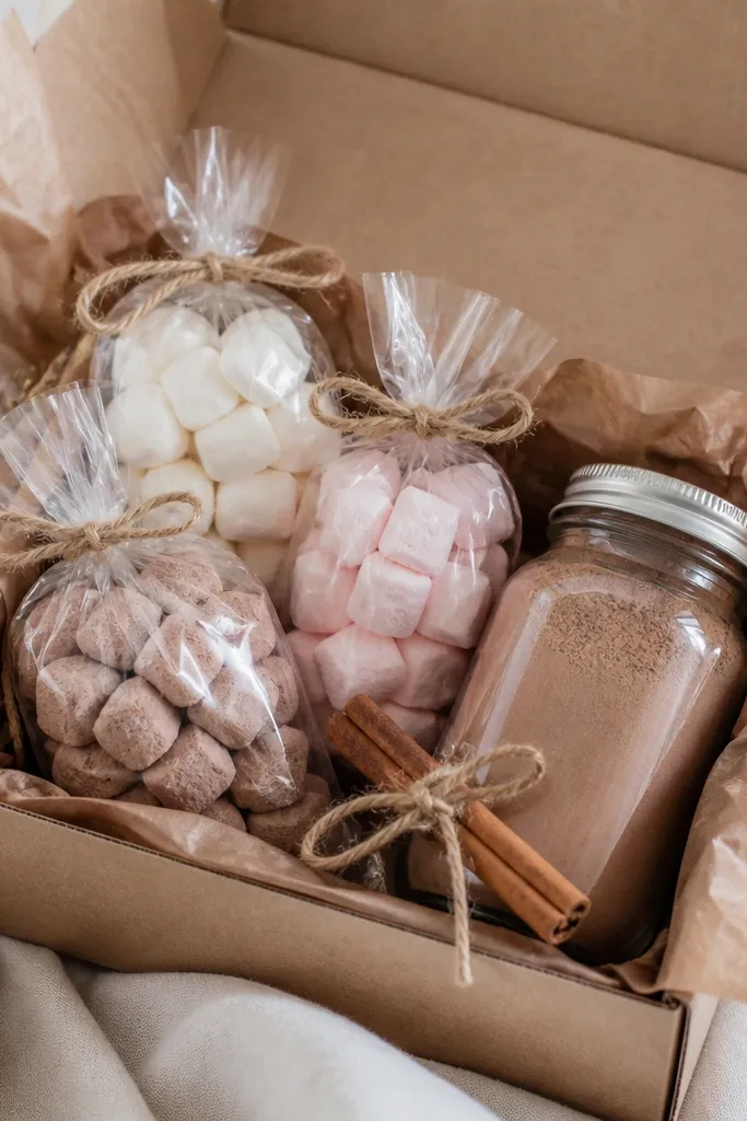 A kraft paper box lined with brown tissue paper holds three small clear bags of marshmallows and a jar of cocoa mix. A cinnamon stick and twine bow sit on top.