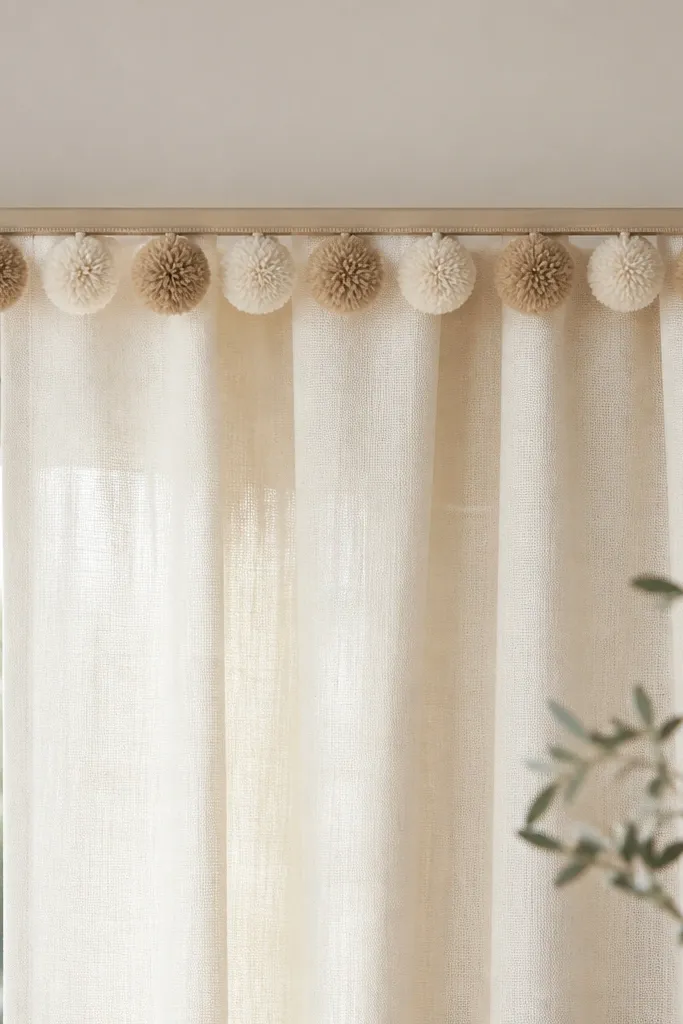 A curtain top border made from yarn pom poms attached in a straight line along a narrow rod or trim piece. The pom poms are in cream and light tan, with small even spacing.