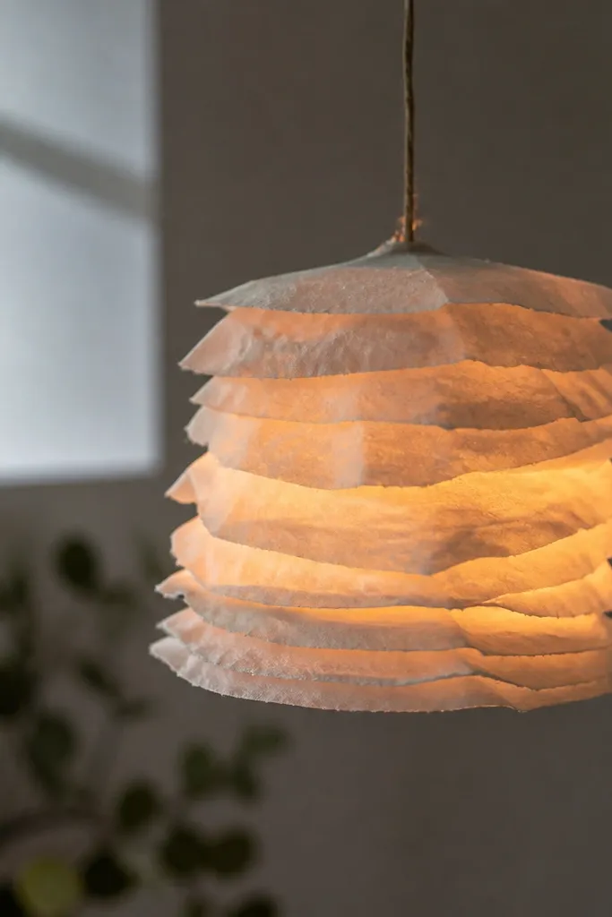 A small hanging paper lantern shade made from layered paper sheets, suspended by string. The lantern glows softly with warm light inside and shows a textured paper surface.