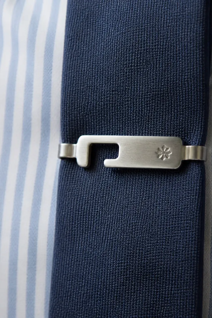 A tie bar made from a repurposed watch strap buckle piece. The metal is brushed and has a small stamped design. Two small clasps clip onto the tie. The tie bar sits on a striped shirt in the photo.