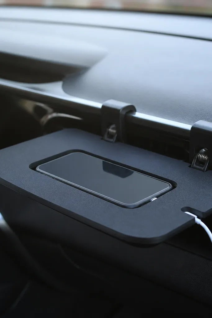 A small curved tray made from coated foam board with a phone-sized cutout. Two spring clips clamp it to an air vent or dashboard edge. A cable passes through a side notch.