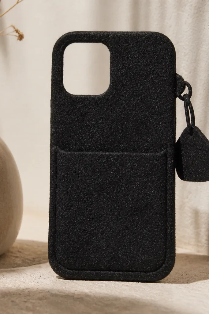 A black phone cover made from thick felt with a shallow front pocket. A small separate charm tab sits on the right side, held by a black elastic loop. The phone's camera area is open, and the felt edges are hand-trimmed with neat, slightly rounded corners.
