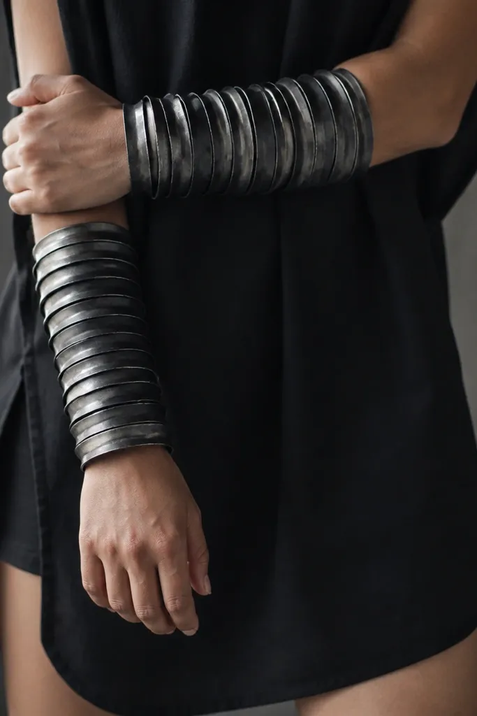 Wrist and forearm armor made from stacked plastic bangles and foil-backed craft strips, painted gunmetal. The wearer has a short black tunic and the armor gleams at the edges.