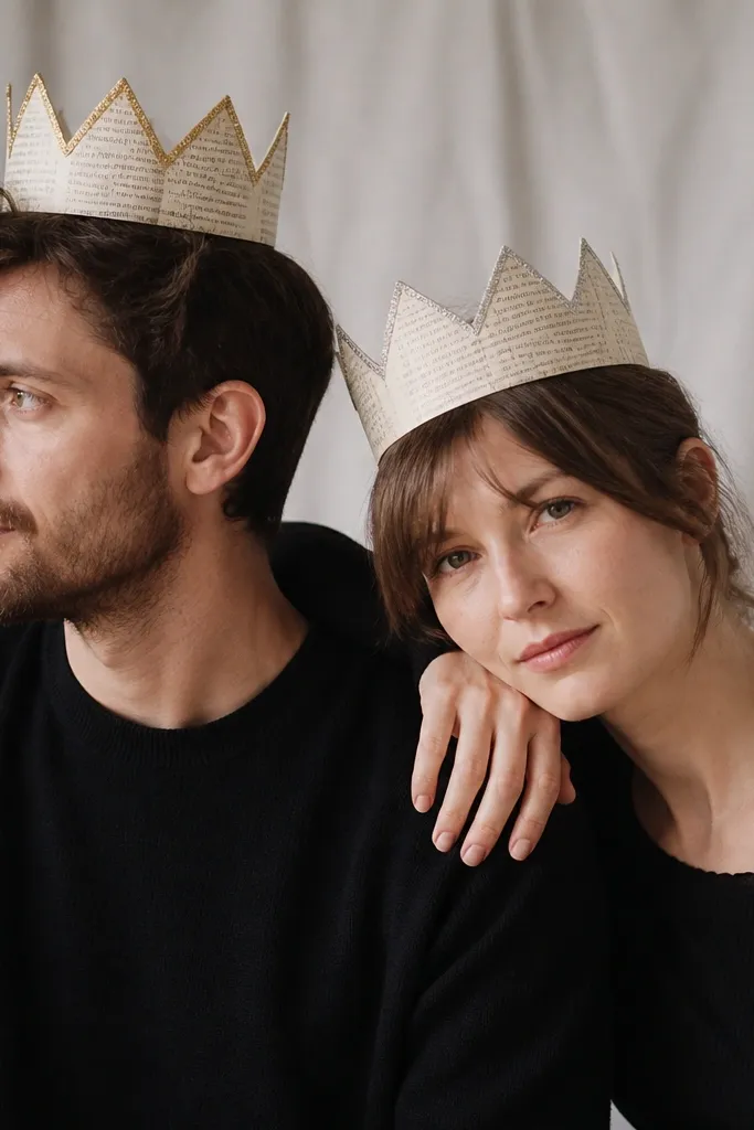 A couple wearing simple black outfits with handmade paper crowns. Each crown is made from the pages of an old paperback, cut into a repeating zigzag top. The king crown has a gold marker border and the queen crown has a silver glitter line.