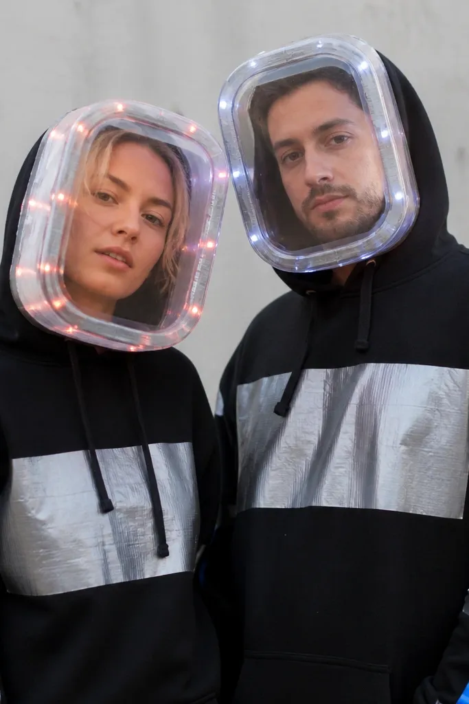 Two people posed together. Each wears a black hoodie with a silver duct-tape "panel" across the chest. Their heads have homemade clear-visor helmets made from a transparent plastic storage lid cut to shape, with small LED puck lights taped around the rim. One partner has a red stripe on the sleeve and the other has a blue stripe, both matching the same silver tape pattern.