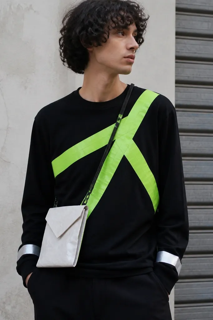 A black long-sleeve shirt with thick neon green duct tape strips forming a diagonal pattern, a small crossbody pouch made from a recycled envelope, and reflective tape bands on the cuffs; the person stands in a street-style pose.