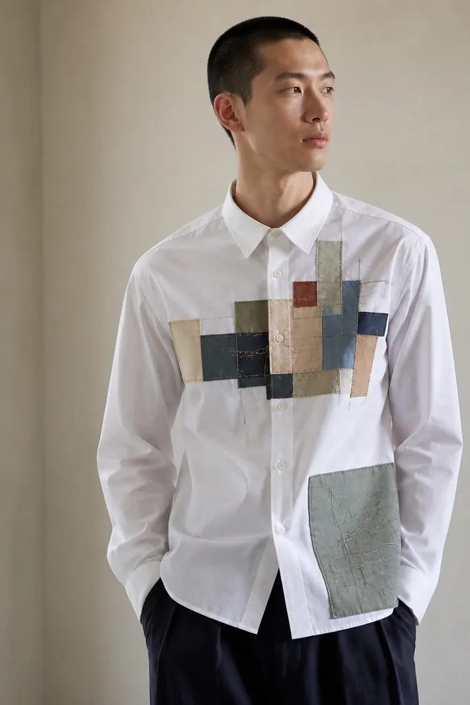 A white button-up shirt with a DIY map-like pattern made from colored fabric rectangles on the chest and one large pocket panel on the hip.