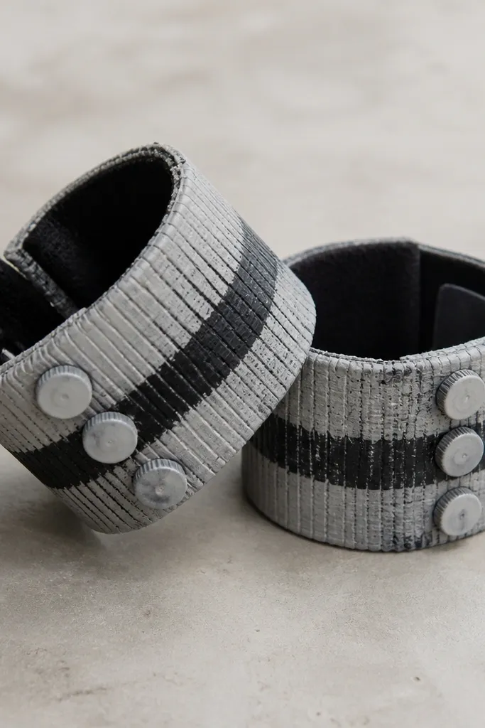 Two chunky arm cuffs made from the sleeves of an old sweater, painted light gray with black stripes. The cuffs have small plastic bottle cap "rivets" and a Velcro closure.