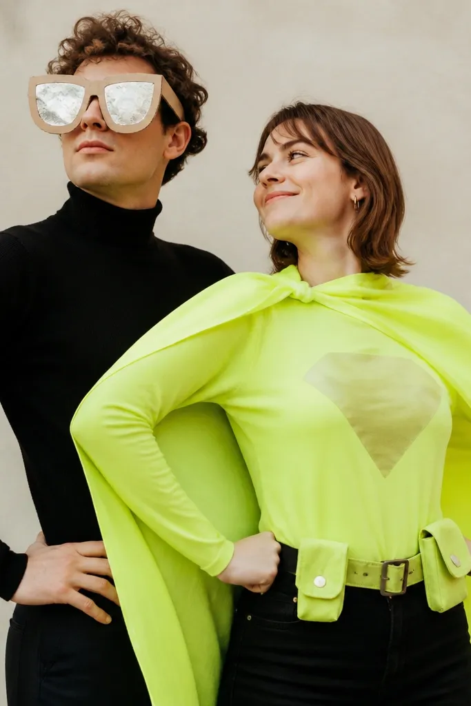 A playful couple: one person wears oversized cardboard sunglasses with reflective paint and a black turtleneck. The other has a bright cape made from neon fabric with a simple emblem on the chest and a mini utility belt.