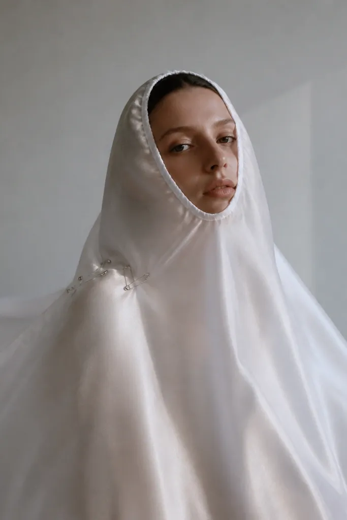 A translucent white satin sheet draped over a person's shoulders. The face opening is shaped like a soft oval with hand-tucked edges. The ghost "sheet" floats slightly, and a few silver safety pins are visible near the shoulder for structure.