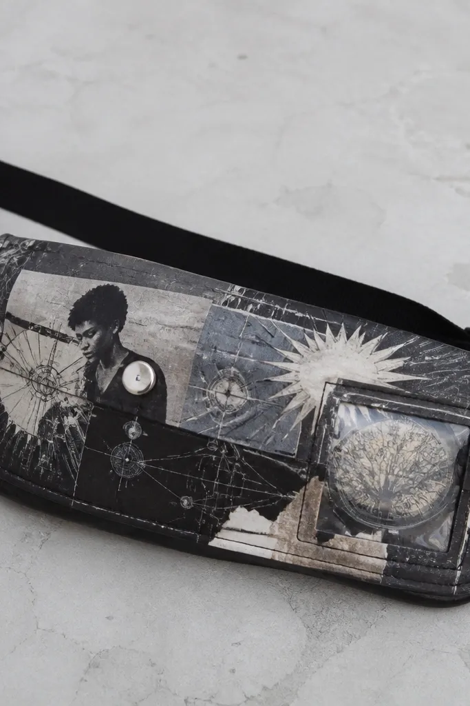 A black belt pack decorated with layered magazine cutouts. The cutouts include tiny technical diagrams and starburst shapes, sealed under clear matte spray. Silver snaps and a small clear plastic window are visible.