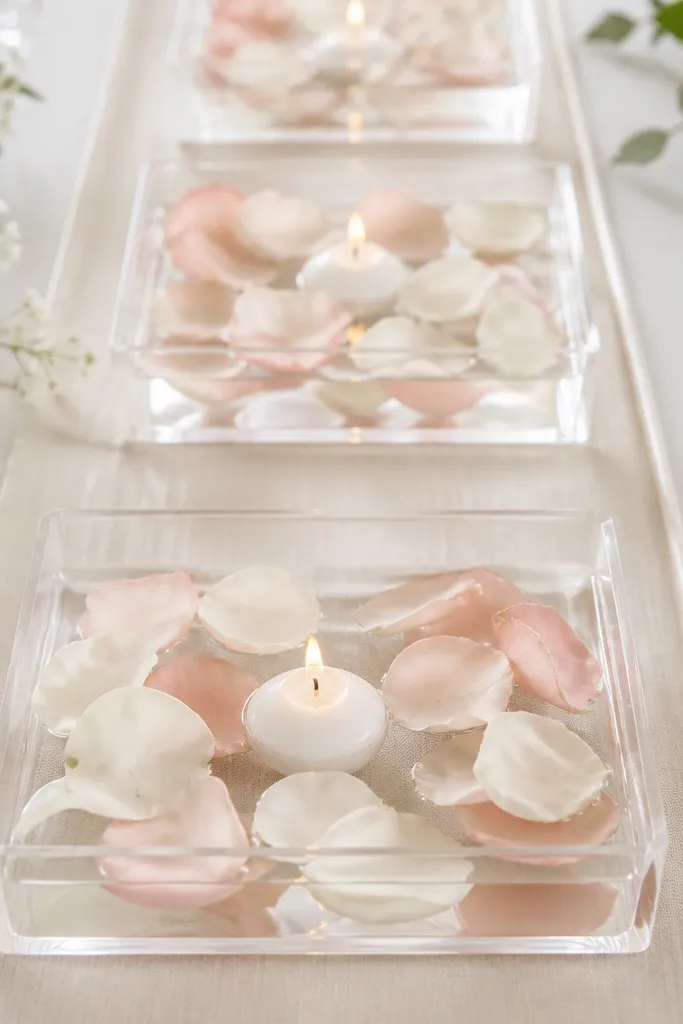 Small clear acrylic trays filled with water. Floating LED candles sit in the middle, surrounded by scattered faux petals in blush and white. The trays are on a table runner.