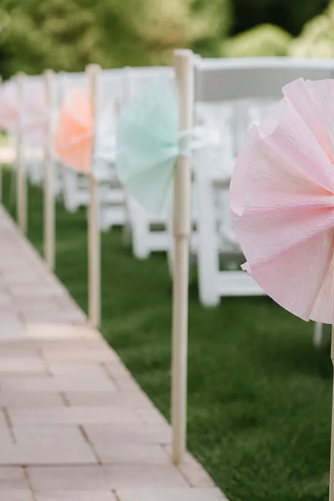 Aisle markers placed along a walkway. Each marker has a small crepe paper shape in pastel colors attached to a ribbon sleeve around a stake. The stakes are spaced evenly.