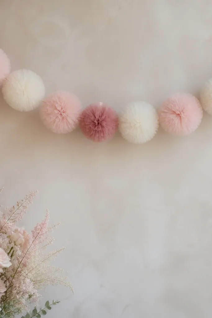 A soft garland made of tulle pom puffs hanging in a gentle curve. Colors include dusty rose, blush pink, and warm white. It's strung on a thin wire line behind a photo backdrop.